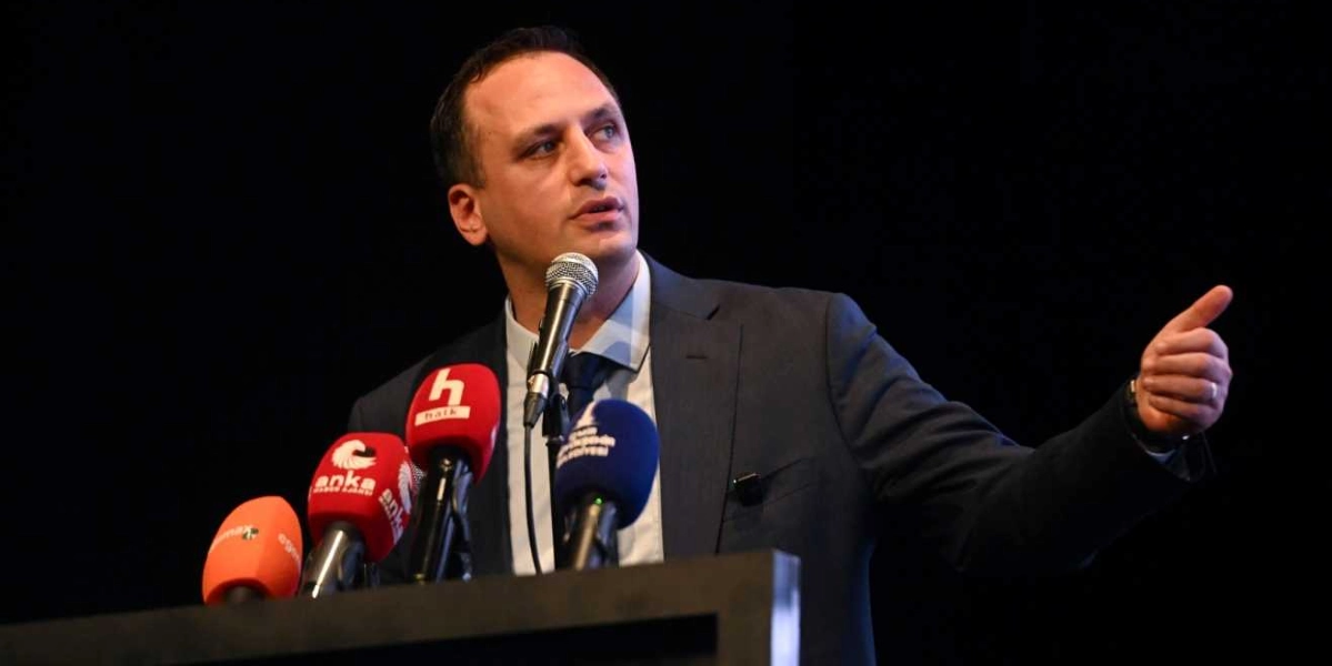 Koyu renkli bir arka plan önünde, kürsüde konuşma yapan bir erkek. Lacivert takım elbiseli ve açık renk gömlekli olan kişi, bir eliyle işaret ederek açıklamalarda bulunuyor. Önündeki mikrofon standında Anka Haber Ajansı, Halk TV ve Bornova Belediyesi logolarının bulunduğu mikrofonlar yer alıyor.