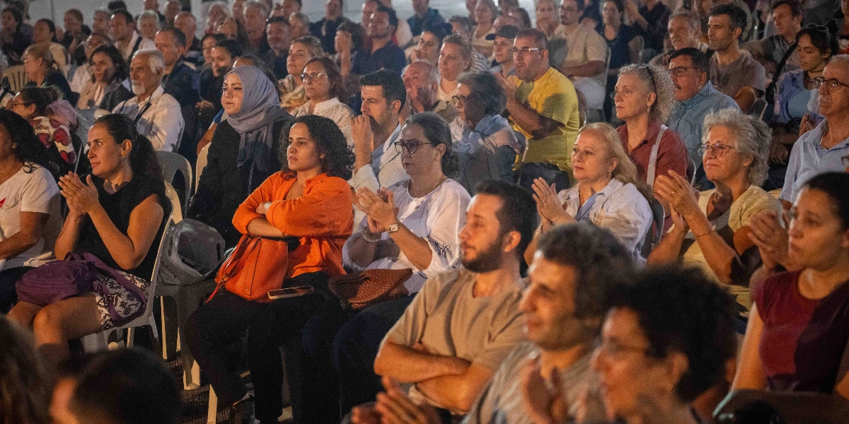 Bornova Kitap Günleri etkinliğinde, plastik sandalyelerde yan yana oturmuş, ilgiyle izleyen ve alkışlayan kalabalık bir izleyici grubu.