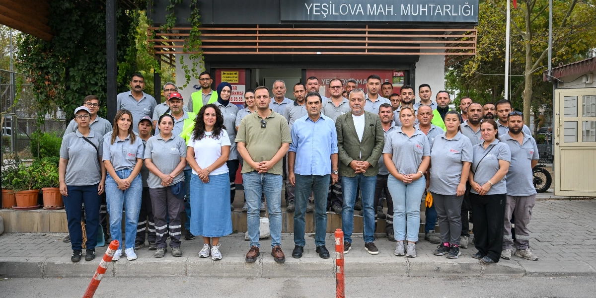 Yeşilova Mahalle Muhtarlığı binasının önünde, Dünya Temizlik Günü kapsamında bir araya gelen, aralarında Bornova Belediyesi çalışanlarının da bulunduğu kalabalık bir grup, hatıra fotoğrafı çektirmek için yan yana duruyor. Ön sırada, merkezde duran iki kişi ve yanlarındaki katılımcılar günlük kıyafetleriyle yer alırken, arka sıralarda bulunan bazı kişiler üzerinde Bornova Belediyesi logolu gri iş tişörtleri ve iş kıyafetleri bulunuyor. Bina girişinin üzerinde 