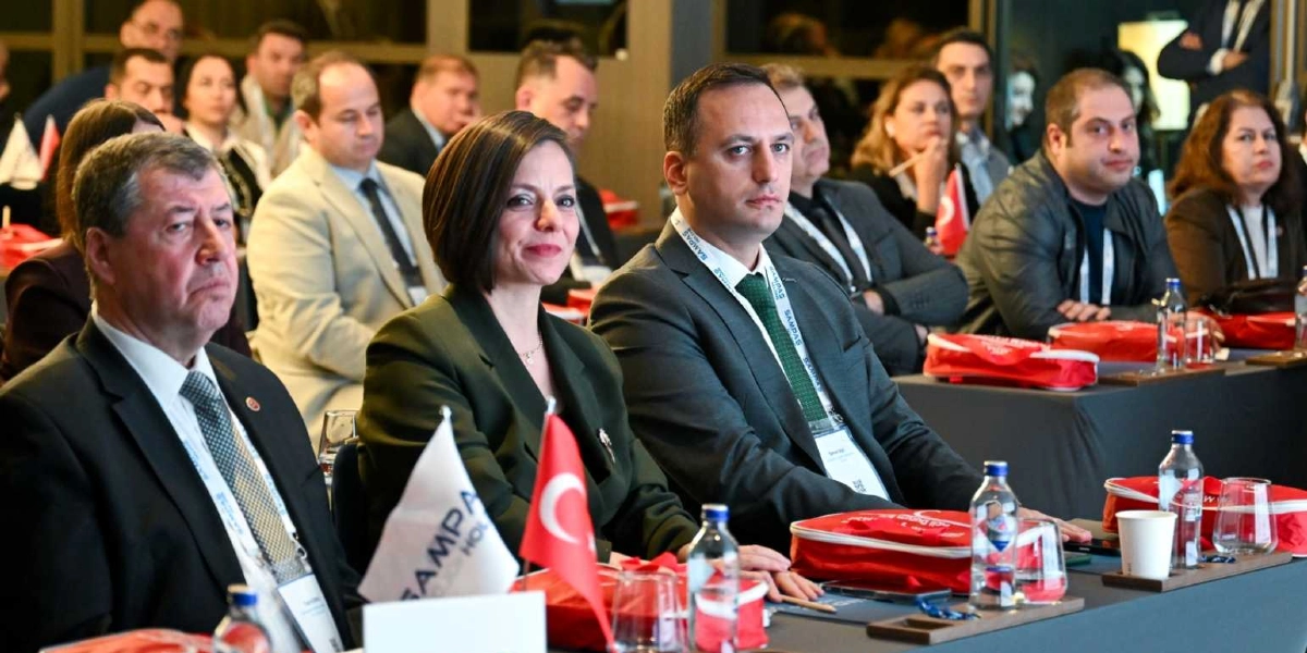 Bir konferans salonunda, ön sırada yan yana oturan katılımcılar yer alıyor. En önde, koyu renk takım elbiseli bir erkek, yanında koyu yeşil ceketli bir kadın ve onun yanında yeşil kravatlı, gri takım elbiseli Başkan Eşki dikkatle karşıya bakıyor. Masaların üzerinde Türk bayrakları, Sampas Holding logolu materyaller, su şişeleri ve katılımcı kartları bulunuyor. Arka planda ise toplantıyı takip eden diğer katılımcılar yer alıyor.