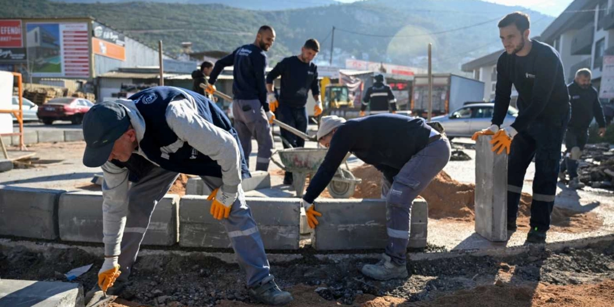 Bornova Belediyesi üniformalı işçiler, yol kenarında bordür taşı döşeme ve çevre düzenleme çalışması yapıyor. Sarı iş eldivenleri takan ekip üyeleri, ellerindeki taşları yerleştirirken ve el arabasıyla malzeme taşırken görülüyor. Arka planda ise araç trafiğinin olduğu bir yol ve binalar yer alıyor.