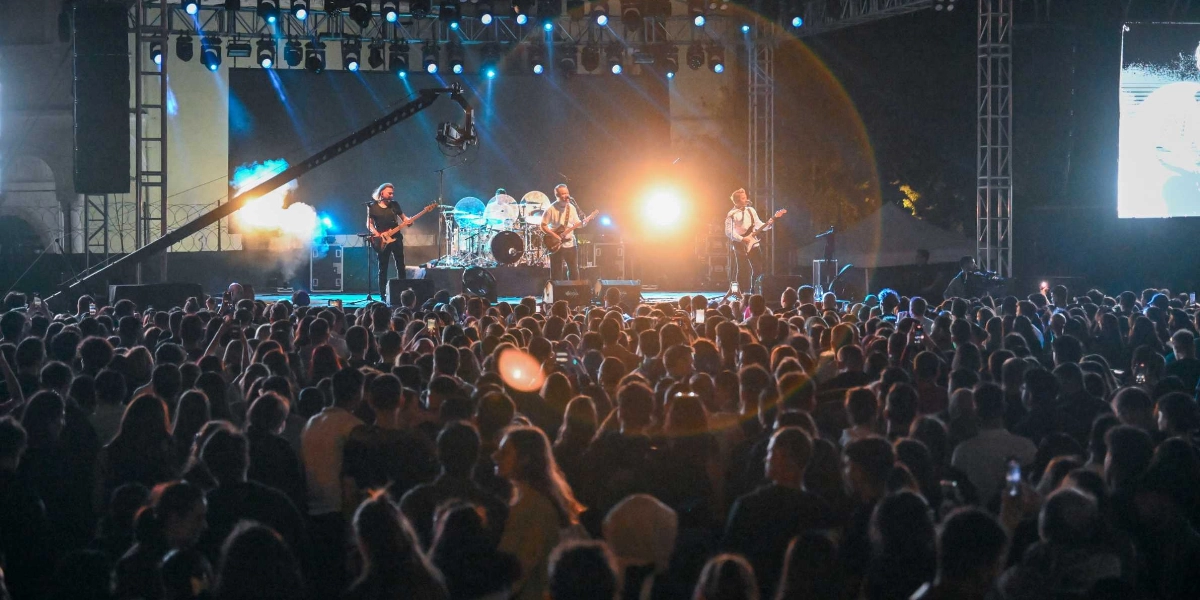 Gece vakti düzenlenen bir açık hava konserinde, sahne ışıkları altında performans sergileyen müzik grubu ve onları izleyen kalabalık bir kitle görülüyor. Sahnedeki grup üyeleri enstrümanlarını çalarken, arkalarındaki dev ekranlarda görüntüler yansıtılıyor ve sahne üzerindeki profesyonel aydınlatma sistemleri ortamı aydınlatıyor. İzleyiciler sahneye doğru dönmüş durumda ve bazıları ellerindeki telefonlarla konseri kaydediyor.