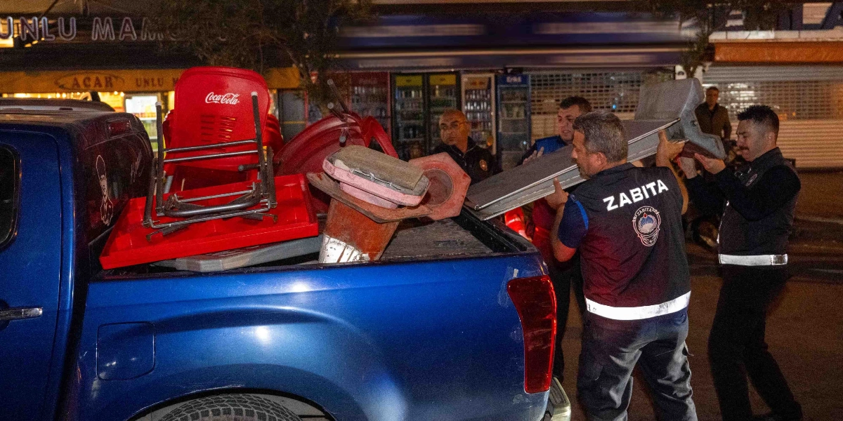 Bornova Belediyesi zabıta ekipleri, gece saatlerinde kaldırım işgali yapan masa, sandalye ve reklam dubası gibi eşyaları toplayarak mavi renkli bir kamyonetin kasasına yüklüyor.