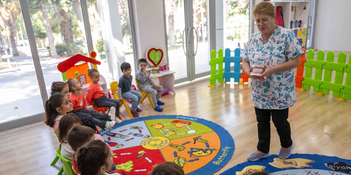 Aydınlık bir anaokulu sınıfında, üzerinde diş desenleri olan beyaz bir forma giymiş kadın eğitmen, elinde tuttuğu büyük bir diş maketiyle çocuklara ağız ve diş sağlığı eğitimi veriyor. Eğitmenin karşısında, renkli bir halının üzerinde oturan küçük bir grup çocuk, dikkatle eğitmeni dinliyor. Sınıfın arka planında oyuncaklar ve çocukların oyun alanını çevreleyen renkli plastik çitler bulunuyor.
