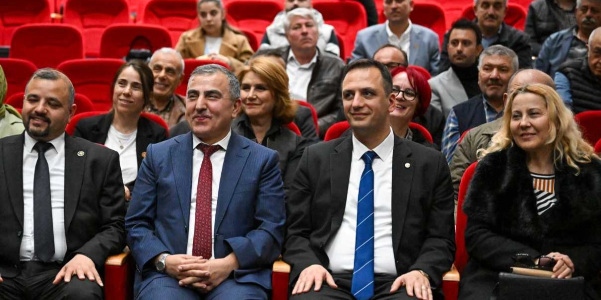 Konferans salonunda kırmızı koltuklarda oturan, aralarında Bornova muhtarlarının da bulunduğu bir grup insan, gülümseyerek objektiflere bakıyor. Ön sırada takım elbiseli ve kravatlı erkekler ile şık giyimli kadınlar yan yana oturuyor.
