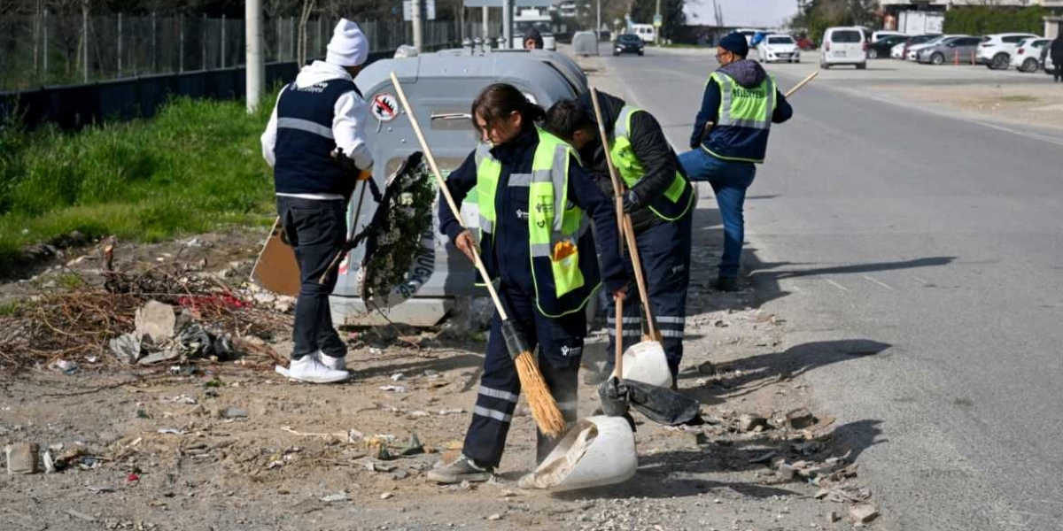 Bornova Belediyesi üniformalı temizlik görevlileri, yol kenarındaki toprak alanda süpürge ve faraşlarla temizlik çalışması yapıyor. Görevliler, büyük bir çöp konteynerinin çevresindeki atıkları toplarken, arka planda park halindeki araçların bulunduğu asfalt bir yol görülüyor.