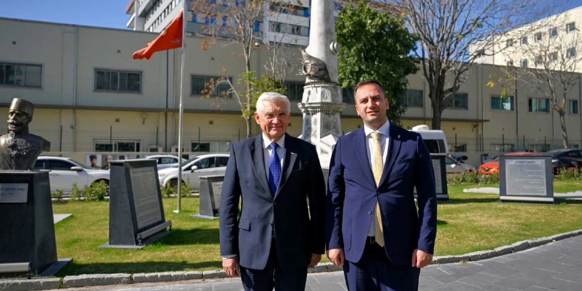 Takım elbiseli iki erkek, açık havada, arkalarında bir anıt ve büstün bulunduğu park benzeri bir alanda yan yana durarak kameraya gülümsüyor. Soldaki kişi beyaz saçlı, lacivert takım elbiseli ve mavi kravatlı; sağdaki kişi ise koyu renk takım elbiseli ve açık sarı desenli kravatlıdır. Arka planda ağaçlar, parktaki bilgilendirme panoları ve park etmiş araçlar yer alıyor.