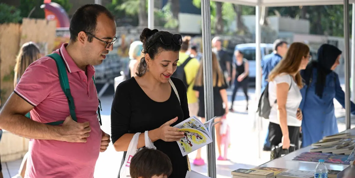 Açık havada kurulan bir kitap fuarı standında, pembe polo yaka tişörtlü ve gözlüklü bir erkek ile siyah bluzlu bir kadının birlikte kitap incelediği an. Kadın elinde tuttuğu resimli bir kitabı dikkatle incelerken, yanındaki erkek ona eşlik ediyor. Arka planda ise stantlar arasında dolaşan diğer ziyaretçiler ve güneşli bir günün atmosferi görülüyor.