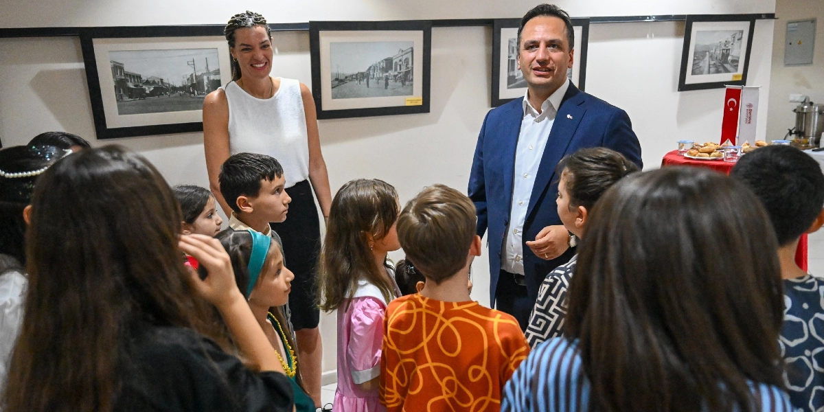 Beyaz bir sergi salonunda, lacivert ceketli bir erkek ve beyaz kolsuz bluz giyen bir kadın, etraflarını saran bir grup çocuğa gülümseyerek bakıyor. Çocuklar sırtları veya profilleri dönük şekilde yetişkinleri dinliyor. Arka plandaki duvarda siyah beyaz eski fotoğrafların yer aldığı çerçeveler asılı duruyor. Sağ taraftaki masada ise üzerinde Türk bayrağı bulunan bir ikram tabağı görülüyor.