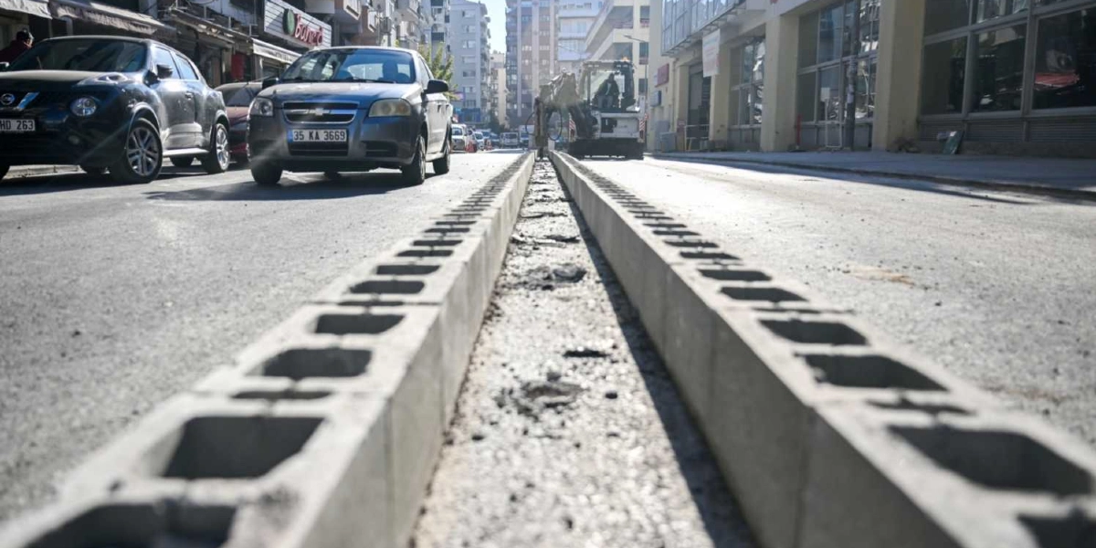 Şehir merkezindeki bir caddede, yolun ortasına boylu boyunca dizilmiş beton briketlerden oluşan bir bariyer hattı görülüyor. Arka planda trafik akışı devam ederken, yolun ilerisinde çalışma yapan bir iş makinesi yer alıyor.