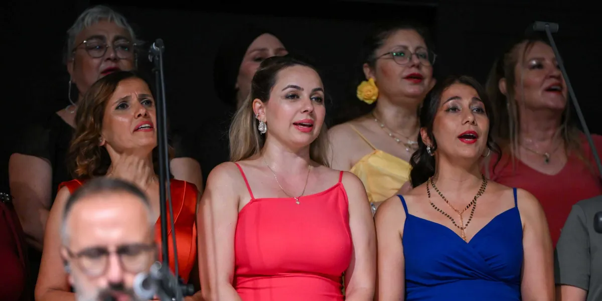 Sahne ışıkları altında türkü söyleyen, farklı renklerde kıyafetler giymiş kadınlardan oluşan bir koro grubu. Ön planda kırmızı ve mavi elbiseli iki kadın şarkı söylerken, arka planda sarı elbiseli ve diğer koro üyeleri onlara eşlik ediyor. En önde ise gözlüklü bir erkek müzisyenin silüeti kısmen görünüyor.
