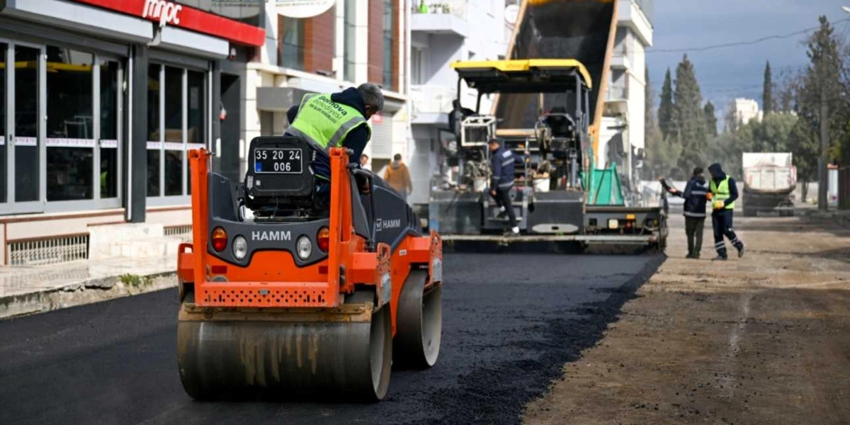 Bornova Belediyesi ekipleri tarafından yürütülen yol asfaltlama çalışmasında, üzerinde Bornova Belediyesi logolu fosforlu yelek bulunan bir operatörün kullandığı turuncu renkli Hamm marka silindir, yeni dökülen asfaltı düzeltiyor. Arka planda ise asfalt serici iş makinesi ve yönlendirme yapan işçiler yer alıyor.