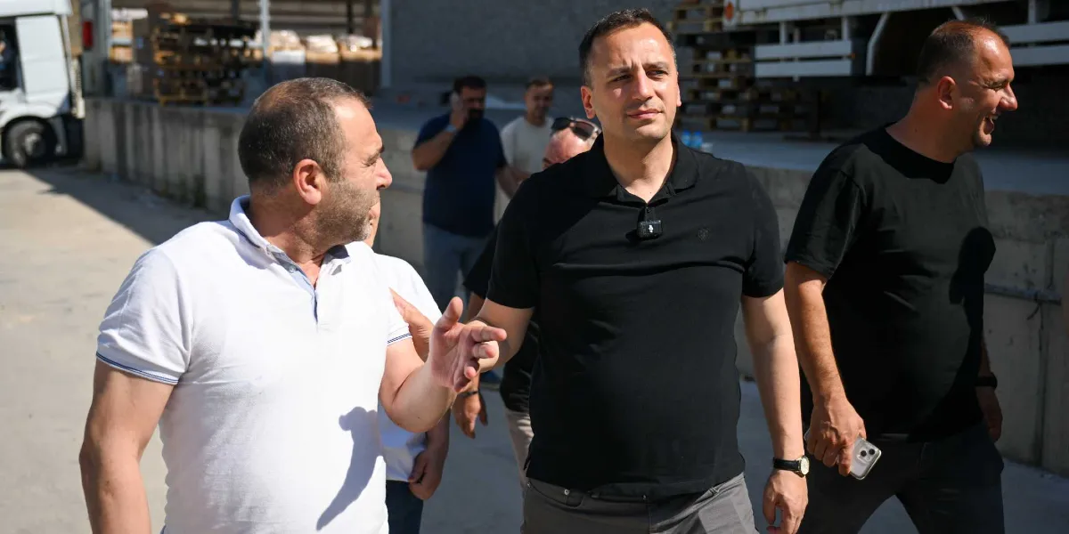 Beyaz polo yaka tişörtlü bir adamın, siyah polo yaka tişörtlü ve yakasında yaka mikrofonu bulunan bir adamla açık havada konuştuğu an. Arka planda ise siyah tişörtlü başka bir adam ve uzakta telefonla konuşan kişiler ile bir kamyonun ön kısmı görülüyor.