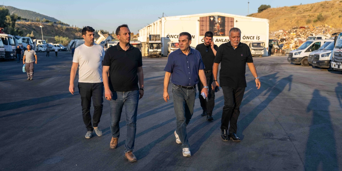 Beş kişilik bir grup erkek, sabah güneşinin altında, arkalarında park halindeki kamyonların ve üzerinde 