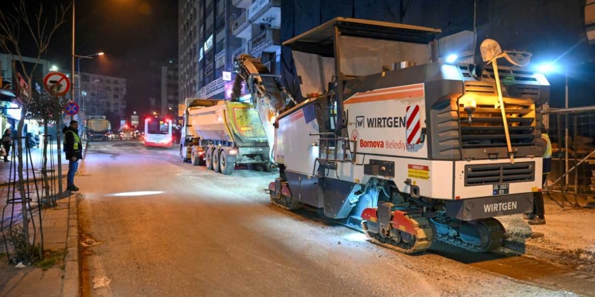 Gece vakti Bornova'da bir cadde üzerinde çalışan Wirtgen marka asfalt kazıma makinesi ve ona bağlı bir hafriyat kamyonu. İş makineleri caddede asfaltı kazırken, çevrede reflektörlü yelek giyen görevliler çalışmayı yönetiyor. Arka planda şehir ışıkları ve trafik akışı görülüyor.