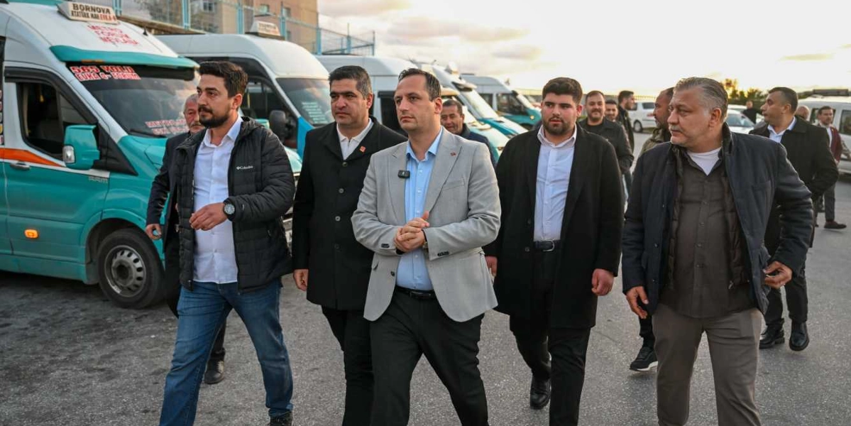 Sabahın erken saatlerinde, arkalarında sıralanmış turkuaz renkli minibüslerin bulunduğu bir alanda yürüyen, çoğunluğu ceketli ve resmi giyimli bir grup erkek.