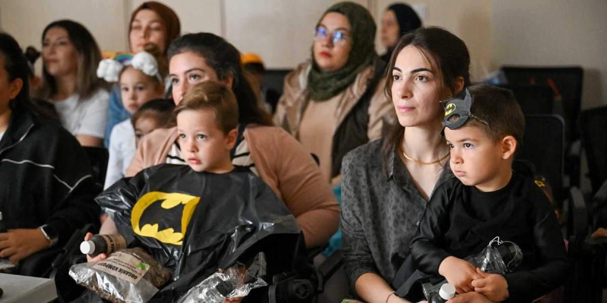 İç mekan bir etkinlikte yan yana oturan, Batman kostümü giymiş iki küçük çocuk ve onlara eşlik eden yetişkinler. Çocuklardan biri üzerinde Batman logosu bulunan siyah bir pelerin giymiş ve elinde 
