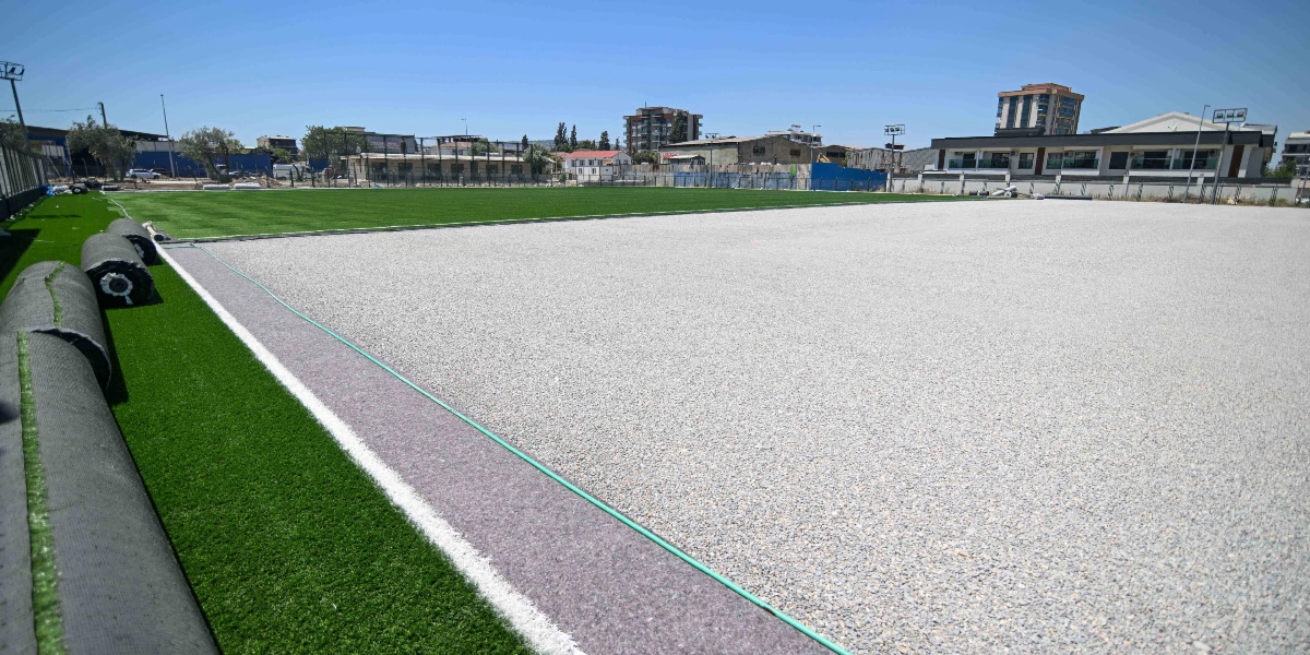 Yapım aşamasındaki bir futbol sahası; zeminin bir kısmı tamamlanmış yeşil suni çimle kaplıyken, diğer geniş bölümü henüz çakıl ve dolgu malzemesiyle kaplı durumda. Sol tarafta serilmeye hazır rulo halindeki suni çimler görülüyor. Arka planda ise şehir yerleşimi ve binalar yer alıyor.