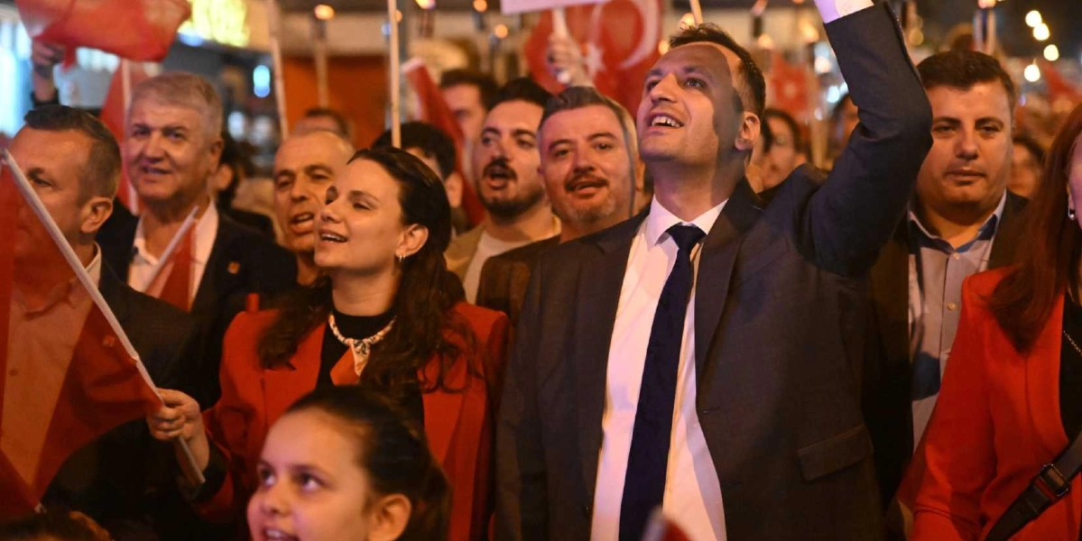 Gece vakti düzenlenen bir yürüyüşte, ellerinde Türk bayrakları taşıyan ve coşkuyla şarkı söyleyen bir grup insan. Ön planda takım elbiseli ve kravatlı bir erkek, elini havaya kaldırmış şekilde heyecanla bir yöne bakıyor; yanında kırmızı ceketli bir kadın ve çevresinde farklı yaşlardan katılımcılar yer alıyor. Kalabalık, 29 Ekim Cumhuriyet Bayramı kutlamaları kapsamında bir arada bulunuyor.