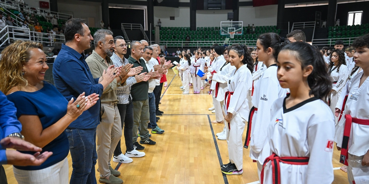 Kapalı bir spor salonunda, beyaz tekvando kıyafetleri ve kırmızı kuşaklarıyla sıralanmış genç sporcuları, bir grup yetişkin ayakta alkışlayarak selamlıyor. Sporcular ve yetişkinler karşılıklı iki sıra halinde duruyor, arka planda ise tribünler ve basketbol potası görülüyor.