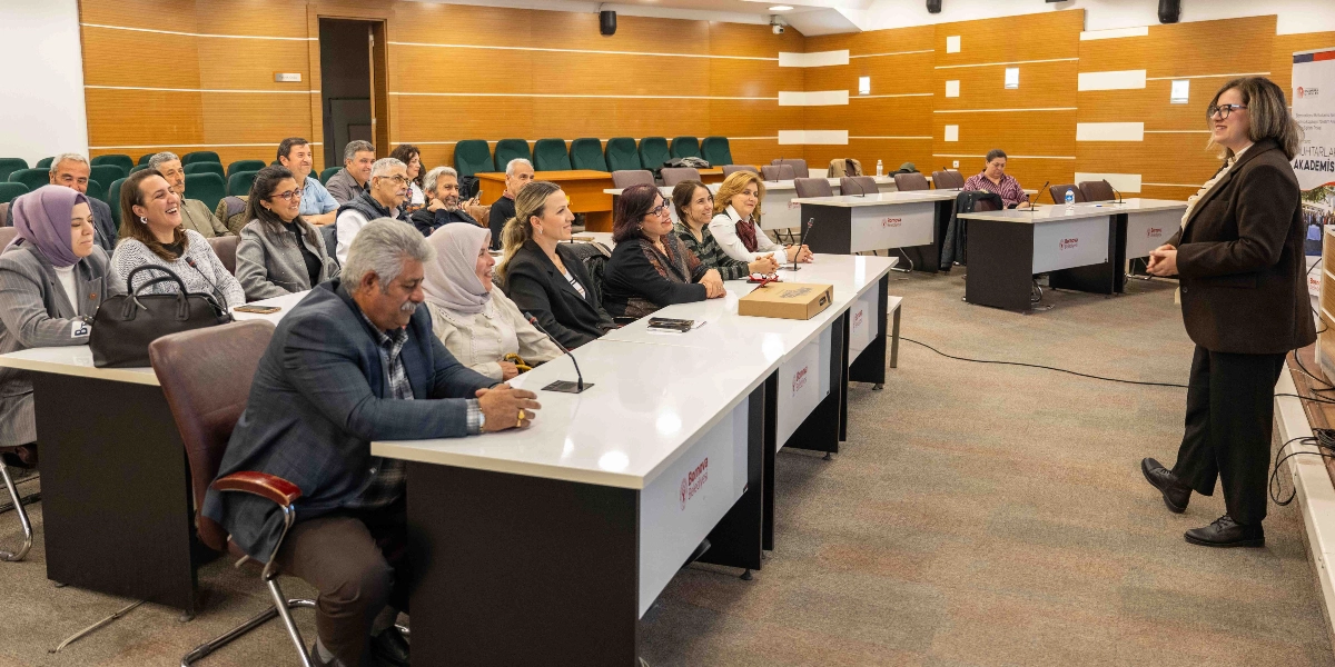 Bornova Belediyesi'nin düzenlediği bir eğitim salonunda, ayakta duran bir eğitmen karşısında oturan muhtarlar grubu görülüyor. Modern bir toplantı salonunda, beyaz masaların etrafında sıralanmış katılımcılar dikkatle dinliyor. Sağ tarafta, üzerinde 