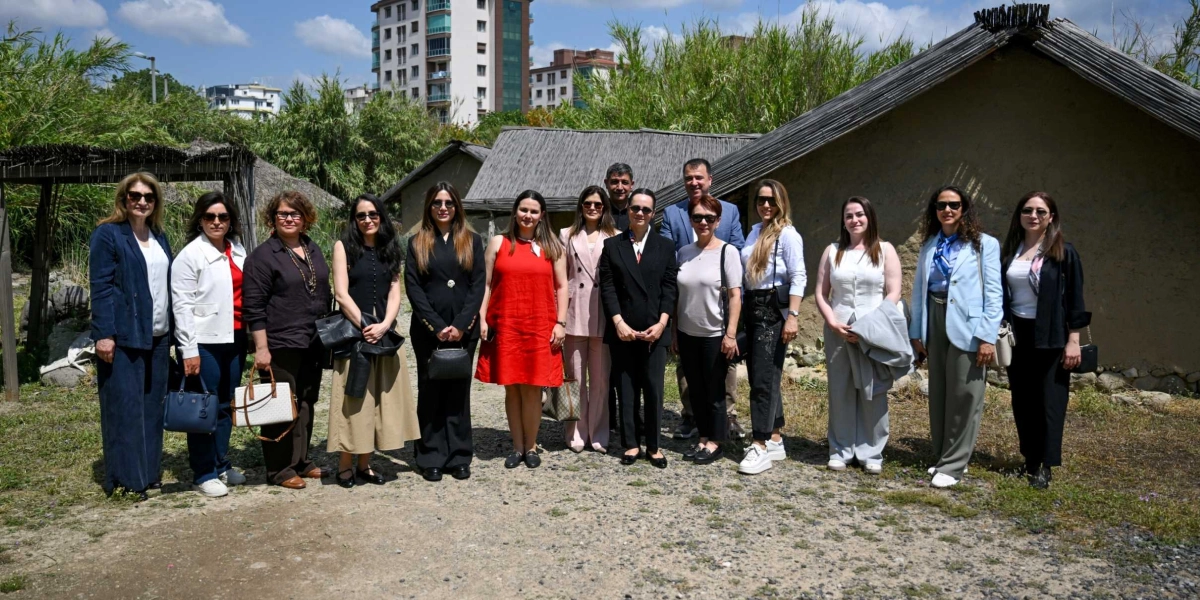 Açık havada, sazdan çatılı geleneksel evlerin önünde yan yana duran, çoğunluğu kadınlardan oluşan bir grup insan. Arka planda yüksek apartmanlar ve yeşil bitki örtüsü görülüyor. Grup, resmi ve günlük kıyafetler içinde, güneşli bir günde objektife gülümsüyor.