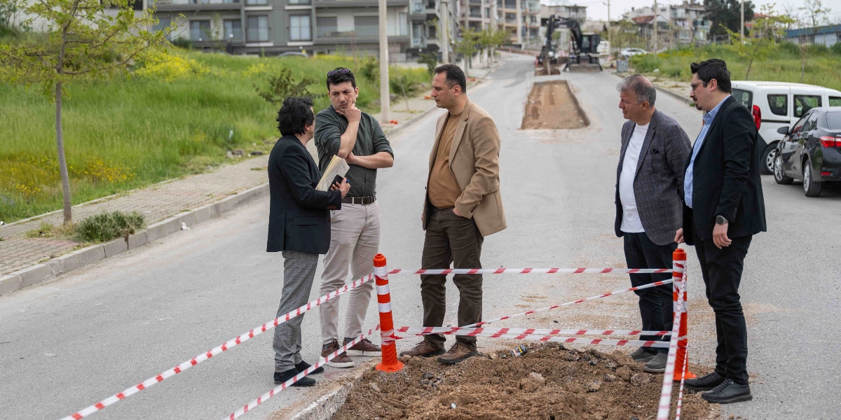 Bornova'da yol çalışması yapılan bir alanda, kırmızı beyaz güvenlik şeridiyle çevrili kazı alanının başında duran beş kişilik bir grup yetkili, devam eden çalışmaları inceleyerek birbirleriyle konuşuyor. Arka planda iş makinesi ve binalar görülüyor.