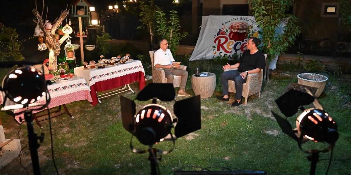 Açık havada, çimenlik bir alanda kurulan çekim setinde iki erkek karşılıklı koltuklarda oturarak sohbet ediyor. Arka planda üzerinde 