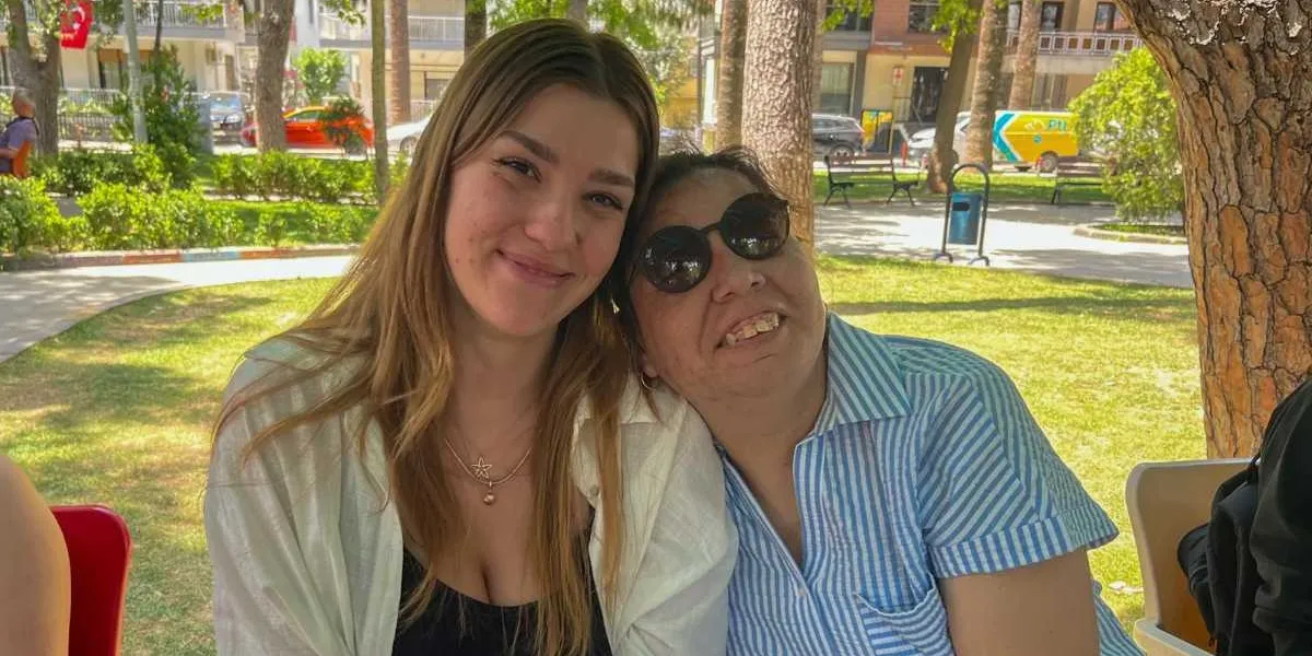 Açık havada, ağaçlık bir parkta yan yana oturmuş iki kadın gülümsüyor. Uzun sarı saçlı genç kadın, siyah bir üst ve üzerine beyaz bir gömlek giymiş; yanında oturan güneş gözlüklü kadın ise mavi beyaz çizgili bir gömlek giyiyor ve başını genç kadının omzuna yaslamış durumda. Arka planda parkın yeşil alanı, ağaçlar ve uzaktan geçen araçlar görülüyor.