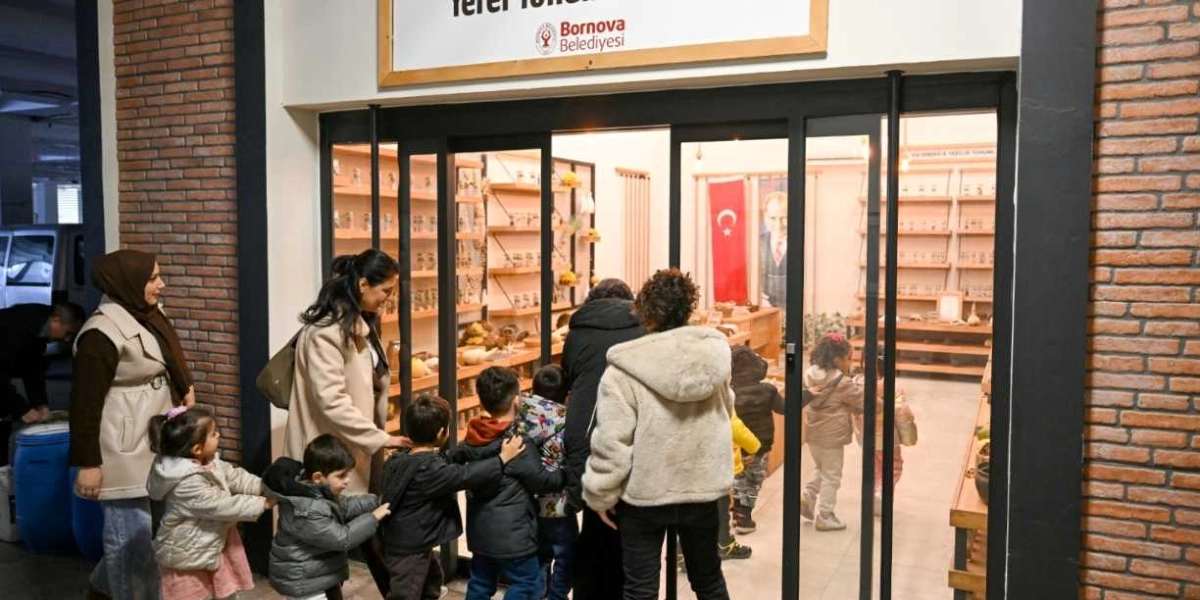 Bornova Belediyesi'ne ait, üzerinde kurum logosunun bulunduğu tabelanın yer aldığı bir dükkanın önünde duran yetişkinler ve çocuklar. Cam kapıdan içerisi görünüyor; içeride raflarda sergilenen ürünler, duvarda asılı bir Türk bayrağı ve Mustafa Kemal Atatürk'ün bir portresi bulunuyor. Çocuklar ve onlara eşlik eden yetişkinler, dükkanın içindeki sergiyi incelemek üzere kapının önünde toplanmış durumda.