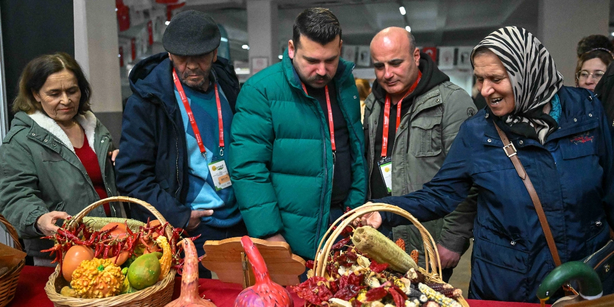 Kapalı bir mekanda düzenlenen etkinlikte, yerel tohumların ve tarım ürünlerinin sergilendiği bir stant başında toplanmış bir grup insan. Önde, başörtülü bir kadın elindeki sepetin içindeki kurutulmuş sebzeleri ve tohumları gösterirken, yanındaki üç erkek ve bir kadın ilgiyle ürünleri inceliyor. Masanın üzerinde hasır sepetler içerisinde kurutulmuş biberler, kabaklar ve çeşitli yerel tarım ürünleri bulunuyor. Katılımcıların boyunlarında Bornova Belediyesi logolu yaka kartları asılı.