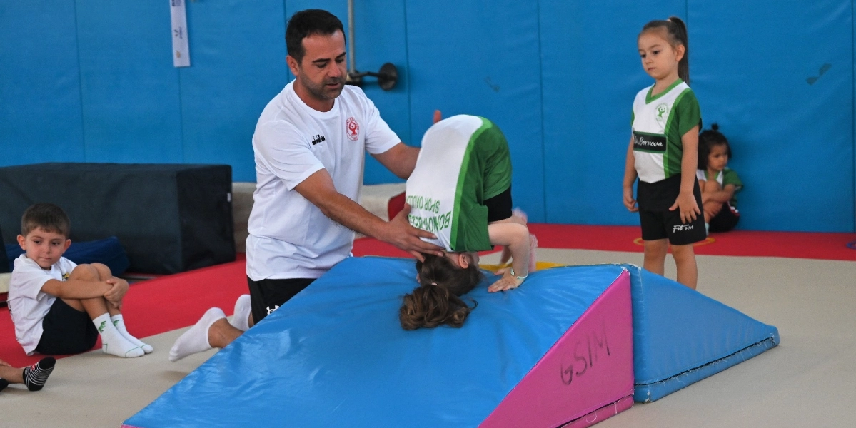 Kapalı bir spor salonunda, beyaz tişörtlü bir eğitmen, mavi ve pembe renkli eğimli bir jimnastik minderinin üzerinde amuda kalkmaya çalışan küçük bir kız çocuğuna destek oluyor. Çocuğun üzerinde üzerinde Bornova Belediyesi Spor Okulları yazan yeşil-beyaz bir forma bulunuyor. Yan tarafta beyaz tişörtlü bir erkek çocuk oturarak izlerken, ayakta bekleyen başka bir kız çocuğu ise çalışmayı takip ediyor. Arka planda mavi duvar kaplamaları yer alıyor.