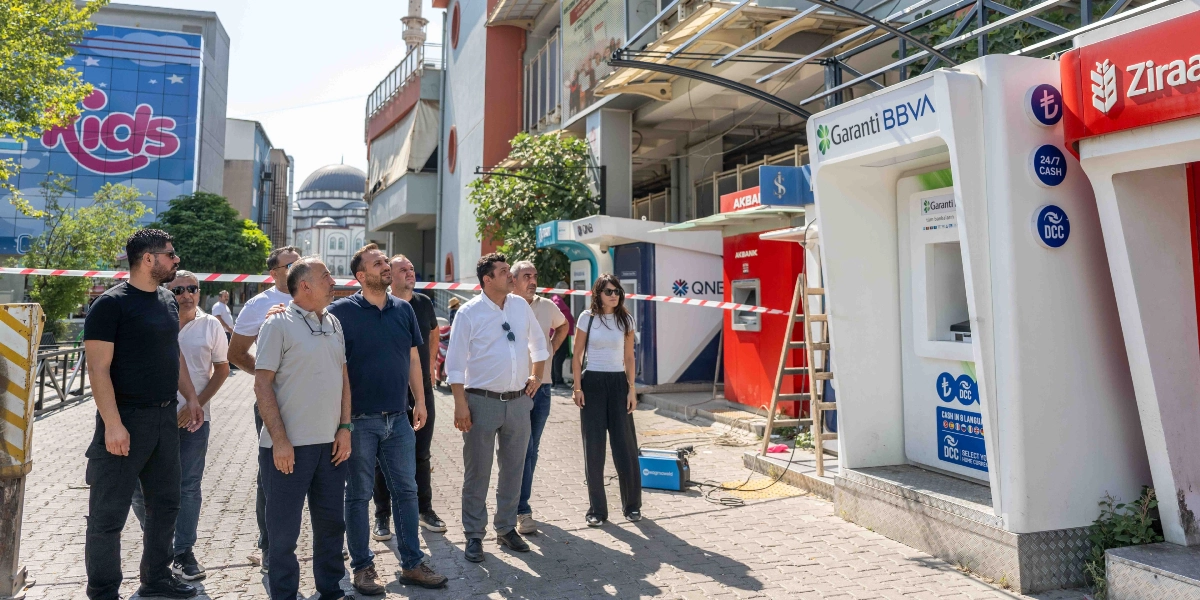 Bornova'da bir grup yetkili, banka ATM'lerinin üzerine yeni monte edilen güneş ve yağmurdan koruyucu sundurma yapısını inceliyor. Arka planda cami ve alışveriş merkezi bulunan açık hava alanında, yan yana dizilmiş Garanti BBVA, Akbank, QNB ve Ziraat Bankası ATM'lerinin önünde duran grup, ATM'lerin üzerini kapatan yeni metal iskeletli koruma sistemine bakıyor.