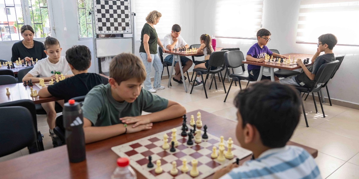 Aydınlık bir sınıfta satranç oynayan çocuklar ve gençler görülüyor. Masalara karşılıklı oturmuş öğrenciler dikkatle hamlelerini yaparken, ayakta duran bir eğitmen öğrencileri gözlemliyor. Odanın köşesinde duvara asılı büyük bir satranç tahtası bulunuyor.