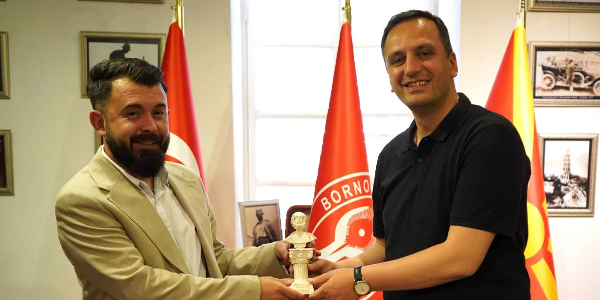 Bej renkli ceket giyen sakallı bir erkek ile siyah polo yaka tişörtlü bir erkek, aralarında küçük bir büst tutarak gülümseyerek poz veriyor. Arka planda Bornova Belediyesi bayrağı ile Makedonya bayrağı yer alıyor, duvarlarda ise çerçeveli eski fotoğraflar bulunuyor.