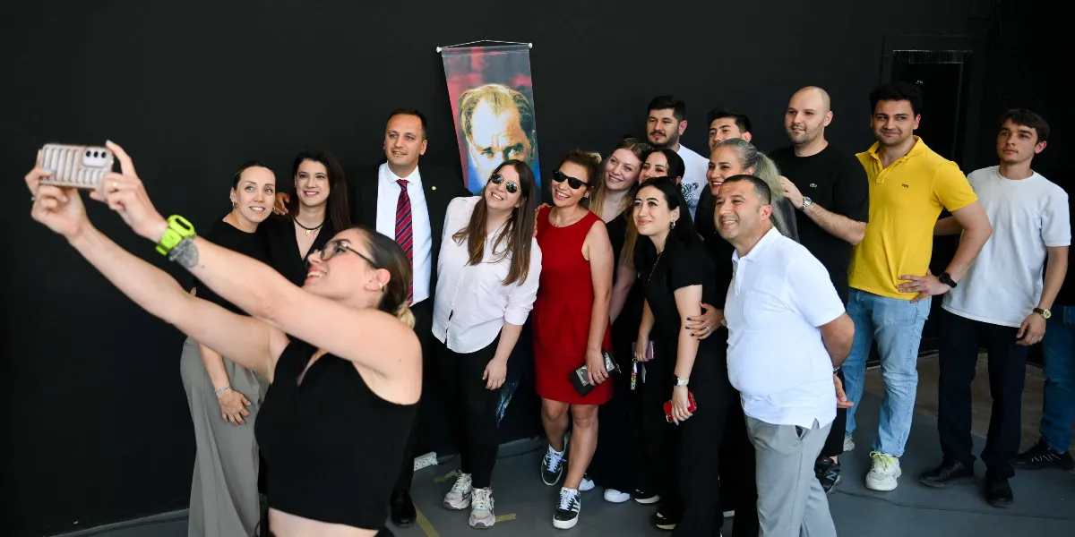 Siyah bir duvarın önünde duran kalabalık bir grup, ön plandaki bir kadının tuttuğu telefonla selfie çekiliyor. Grubun arkasında Mustafa Kemal Atatürk'ün portresinin yer aldığı dikey bir afiş bulunuyor. Farklı kıyafetler içindeki grup üyeleri gülümseyerek kameraya bakıyor.
