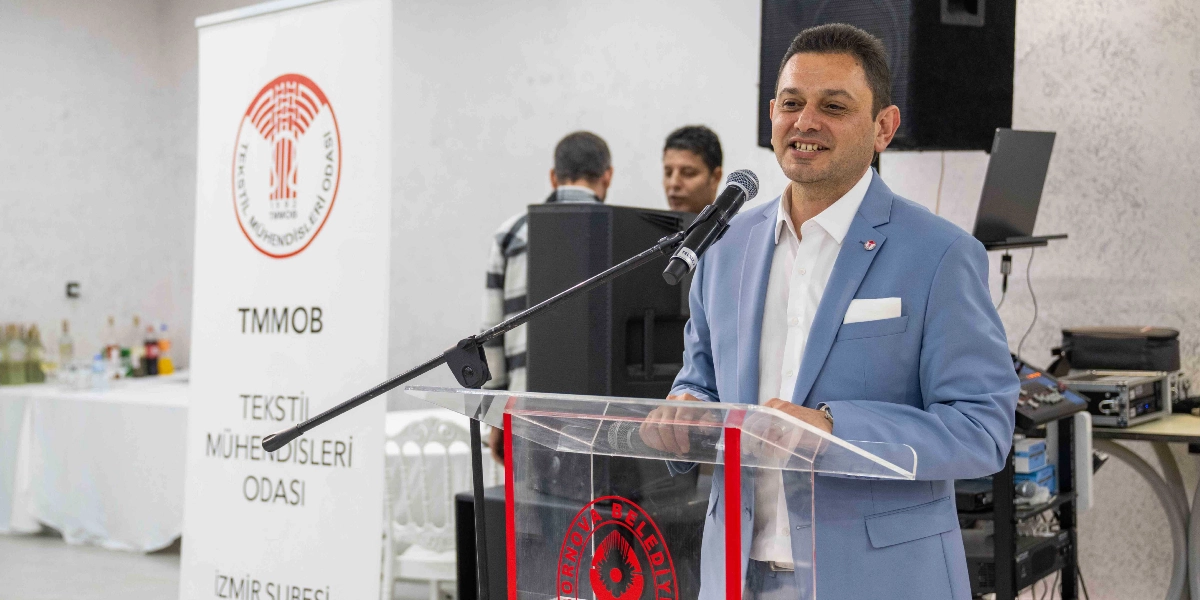 Mavi ceketli ve beyaz gömlekli bir erkek, Bornova Belediyesi logolu şeffaf bir kürsüde mikrofon başında konuşma yapıyor. Arka planda üzerinde TMMOB Tekstil Mühendisleri Odası İzmir Şubesi yazan dikey bir afiş ve etkinlik alanında bulunan diğer katılımcılar yer alıyor.