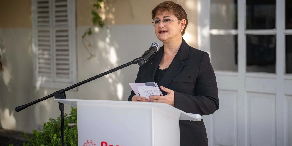 Kısa kahverengi saçlı, gözlüklü ve siyah ceketli bir kadın, dış mekanda beyaz bir kürsü arkasında mikrofonla konuşma yapıyor. Elinde üzerinde notlar bulunan küçük bir kağıt tutuyor. Kürsünün ön kısmında kısmen görünen kırmızı logolu bir yazı yer alıyor. Arka planda beyaz panjurlu pencereler ve yeşil bitkiler bulunuyor.