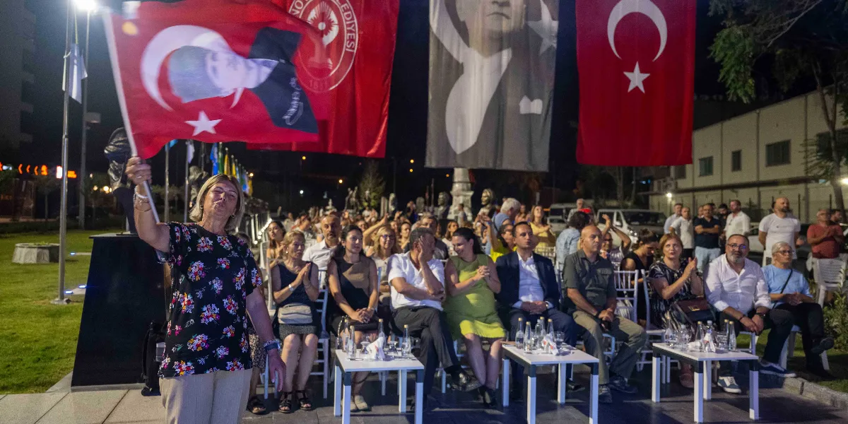 Gece vakti açık havada düzenlenen bir etkinlikte, çiçek desenli bluz giymiş bir kadın elinde üzerinde Atatürk portresi bulunan Türk bayrağını sallıyor. Arka planda Bornova Belediyesi logosunun yer aldığı kırmızı bir bayrak, büyük bir Atatürk posteri ve Türk bayrakları asılı. İzleyiciler, sandalyelerde oturarak etkinliği takip ediyor ve alkışlıyor.