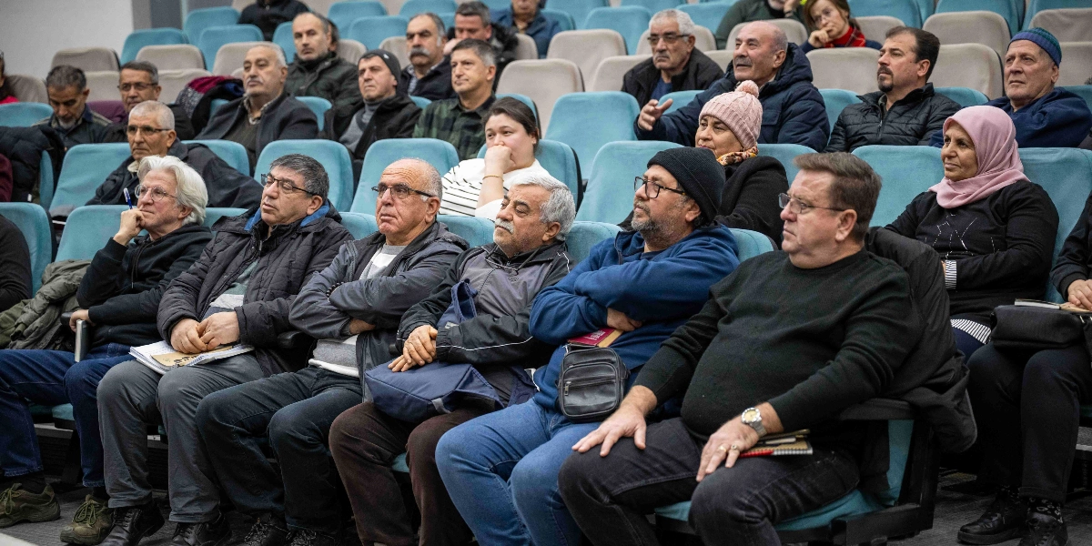 Konferans salonunda, turkuaz renkli koltuklarda oturan ve dikkatle dinleyen, farklı yaş gruplarından oluşan kalabalık bir katılımcı grubu.