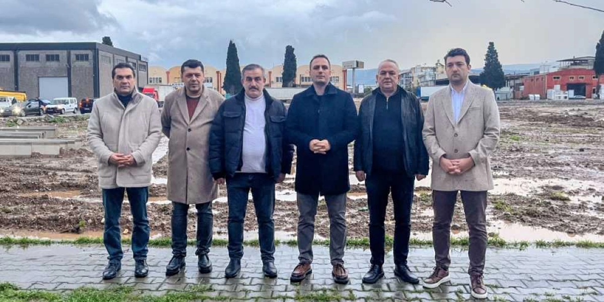 Açık havada, çamurlu ve boş bir arazi önünde yan yana duran, kışlık kıyafetler giymiş altı erkek. Arka planda sanayi tipi yapılar ve ağaçlar görülüyor.