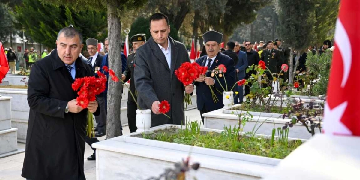 Açık havada, mermer mezarların bulunduğu bir şehitlikte, resmi kıyafetli kişiler ellerindeki kırmızı çiçekleri mezarlara bırakıyor. Ön planda koyu renk palto giymiş üç erkek, ellerindeki kırmızı çiçek demetlerini mezarların üzerine yerleştirirken görülüyor. Arka planda ise askeri üniformalı ve resmi kıyafetli diğer katılımcılar, ellerinde Türk bayrakları ile anma törenine katılıyor.