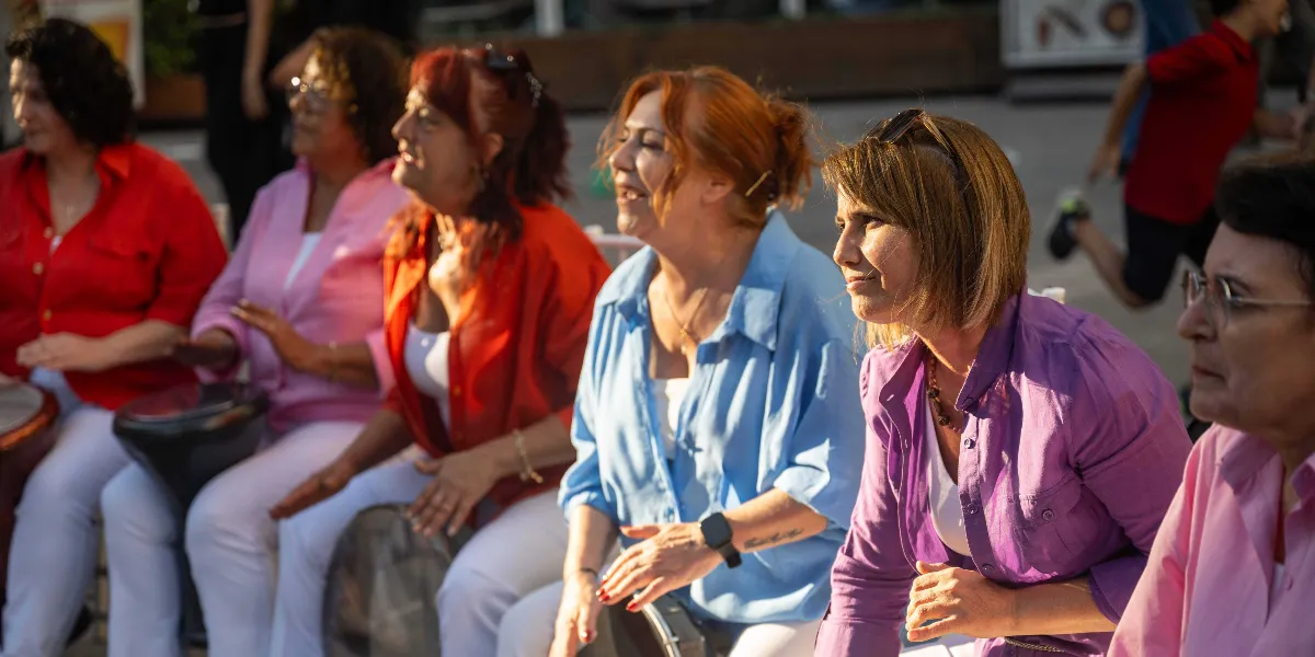 Açık havada yan yana oturmuş, renkli gömlekler giyen ve önlerindeki darbuka benzeri vurmalı çalgıları çalan bir grup kadın. Kadınlar neşeli bir ifadeyle bir yöne doğru bakıyorlar; aralarında kırmızı, pembe, mavi ve mor tonlarında kıyafetler giyenler bulunuyor. Arka planda ise hareket halindeki bir kişinin silüeti seçiliyor.