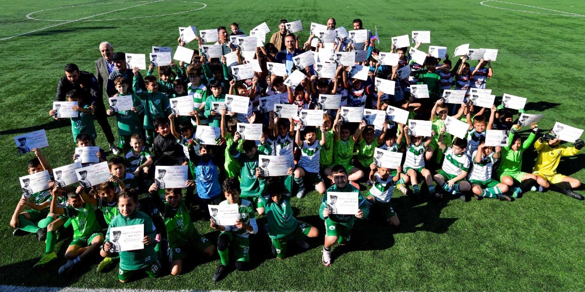 Yeşil bir futbol sahasında, üzerinde Atatürk fotoğrafı bulunan katılım belgelerini havaya kaldırarak poz veren çok sayıda çocuk ve onlara eşlik eden yetişkinler. Çocukların çoğu yeşil-beyaz futbol formaları giymiş durumda ve hepsi neşeli bir şekilde kameraya gülümsüyor.
