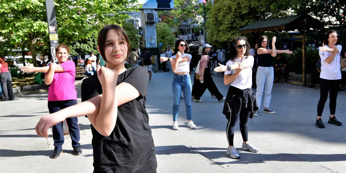 Açık havada, park benzeri bir alanda bir araya gelen kadınlar, eğitmenleri eşliğinde kollarını esnetme hareketleri yapıyor. Ön planda siyah tişörtlü genç bir kızın odak noktasında olduğu grupta, farklı yaşlardan kadınlar spor kıyafetleriyle yan yana dizilmiş şekilde egzersiz hareketlerini uyguluyor. Arka planda ise günlük kıyafetleriyle yürüyen insanlar ve ağaçlıklı bir meydan görünümü yer alıyor.
