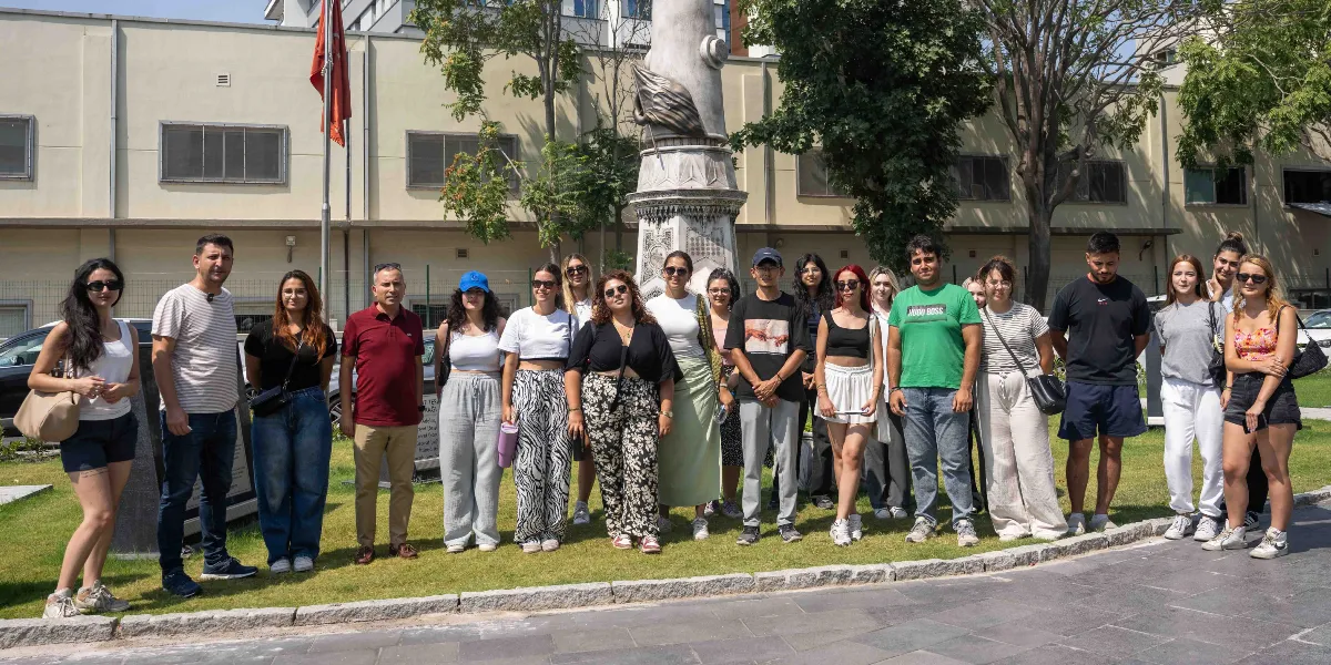 Abide-i Hürriyet Meydanı'ndaki anıtın önünde yan yana dizilmiş, günlük kıyafetler içindeki gençlerden ve yetişkinlerden oluşan yaklaşık yirmi kişilik bir grup, güneşli bir günde poz veriyor. Arka planda açık renkli, uzun bir bina ve yeşil ağaçlar yer alıyor.