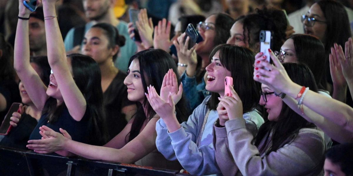 Bir konser alanında sahneye doğru heyecanla bakan, alkışlayan ve telefonlarıyla görüntü kaydeden gençlerden oluşan kalabalık bir grup.