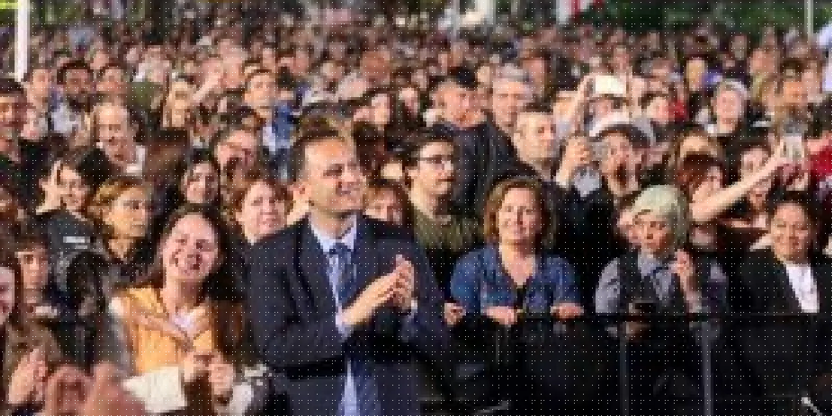 Açık havada düzenlenen bir etkinlikte, ön planda takım elbiseli bir erkek alkışlayarak gülümserken, arkasında onu izleyen ve bazıları telefonlarıyla görüntü alan kalabalık bir insan grubu yer alıyor.