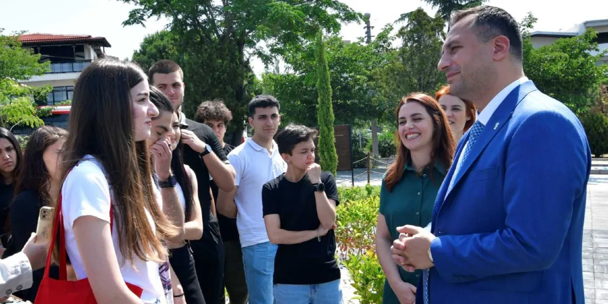 Mavi ceketli bir yetkili, dış mekanda bir grup genç öğrenci ve bir kadın eğitmenle sohbet ediyor. Öğrenciler dikkatle dinlerken, eğitmen gülümseyerek konuşmaya eşlik ediyor. Arka planda ağaçlar ve park benzeri bir alan görülüyor.