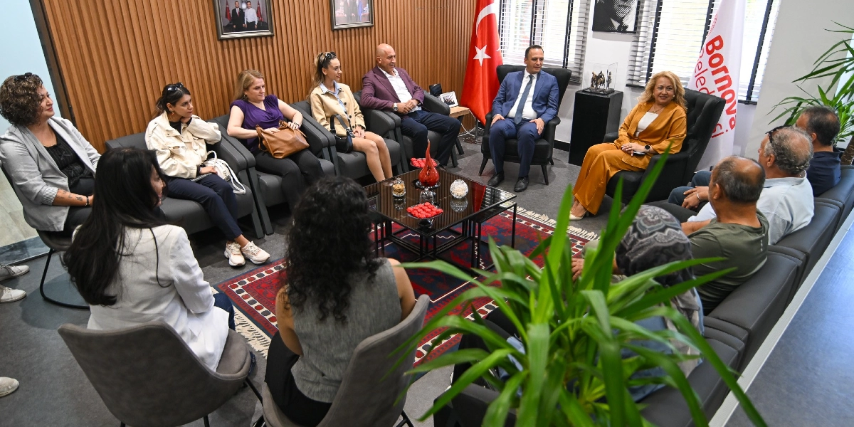 Modern bir ofis ortamında, Bornova Belediyesi yetkililerinin ve vatandaşların katılımıyla gerçekleşen bir toplantı. Odanın merkezinde yer alan sehpanın etrafında, siyah deri koltuklarda oturan yaklaşık 15 kişilik bir grup birbirleriyle sohbet ediyor. Arka planda Türk bayrağı ve Bornova Belediyesi flaması bulunurken, duvarlarda çerçeveli fotoğraflar asılı. Katılımcılar arasında takım elbiseli bir erkek ve hardal sarısı kıyafetli bir kadın dikkat çekiyor; ortamda resmi ancak samimi bir dayanışma havası hakim.